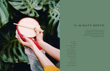 Load image into Gallery viewer, Person holding a red and beige bento box with chopsticks, surrounded by green leaves. | Hadley Green Home
