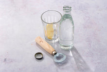 Load image into Gallery viewer, Clear glass bottle, glass with a lemon wedge, wooden bottle opener, and bottle cap on a light gray surface. | Hadley Green Home