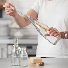 Load image into Gallery viewer, A person in a white shirt holds a long-handled Eco Living glass bottle brush inside a clear glass bottle. Two smaller empty glass bottles and a bar of soap sit on a countertop in a softly blurred kitchen setting. | Hadley Green Home