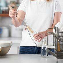Load image into Gallery viewer, A person in a white t-shirt and blue jeans holds a glass bottle with a swing-top closure while cleaning it with a very narrow Eco Living bottle brush. The scene is set in a bright kitchen with a dish rack and bowls visible | Hadley Green Home